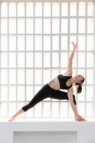 Eine junge athletische Frau in schwarzer Sportbekleidung praktiziert neben einem großen Fenster in ihrem Studio Yoga. Raum für Text. - Foto, Bild