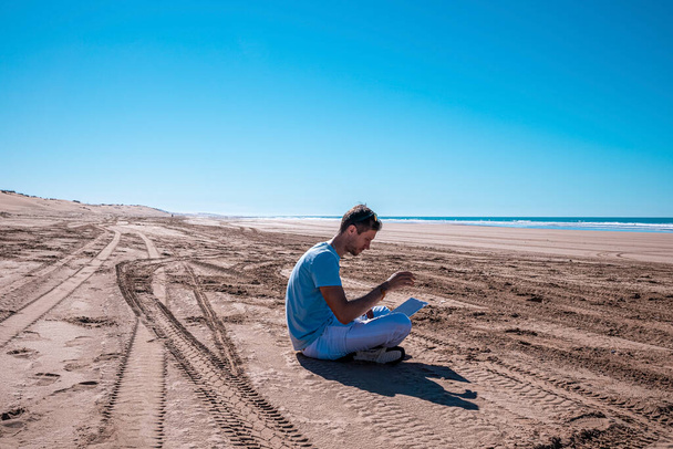 Young man working on laptop computer sitting on sand with tire track prints and seascape against sky - Foto, Bild