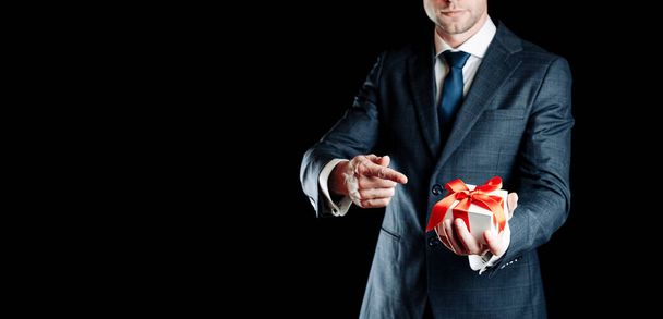 Άνθρωπος δώρο. Ευτυχισμένος νεαρός επιχειρηματίας κρατώντας δώρο έκπληξη κουτί δώρο με κόκκινη κορδέλα απομονώνονται σε μαύρο φόντο. Μαύρη Παρασκευή, Ημέρα του Αγίου Βαλεντίνου συγχαρητήρια πανό - Φωτογραφία, εικόνα