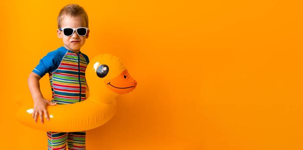 バナー幸せな小さな笑顔の子供の水泳スポーツ少年は、太陽のメガネの水着を着用し、子供インフレータブルスイミングリング黄色のアヒルのポイントを離れて、孤立オレンジの背景に。暑い夏休みコピースペース - 写真・画像