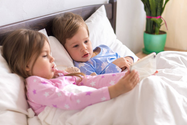 I bambini piccoli di età prescolare fratelli minori guardano insieme i cartoni animati utilizzano il dispositivo smartphone. Bambino in pigiama sul letto bianco a casa camera da letto. Famiglia, Tempo libero, Infanzia, Amicizia - Foto, immagini