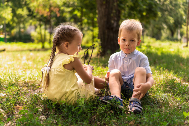 Двоє щасливих усміхнених веселих малюків дошкільного віку близнюки брати і сестри діти брат сестра хлопчик дівчина сидить разом на газоні на газоні в парку в сонячну спекотну літню погоду. дитинство, дружба, концепція сім'ї
 - Фото, зображення