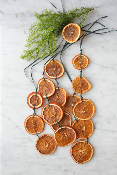 Χειροποίητες Χριστουγεννιάτικες γιρλάντες από αποξηραμένες φέτες πορτοκαλιού σε μαρμάρινη επιφάνεια, τραβηγμένες ακριβώς από πάνω - Φωτογραφία, εικόνα
