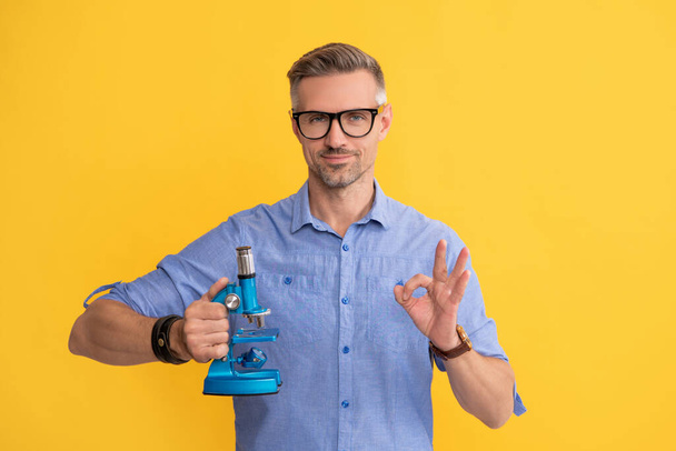smiling scientist man in glasses hold microscope show ok gesture on yellow background, science - Φωτογραφία, εικόνα