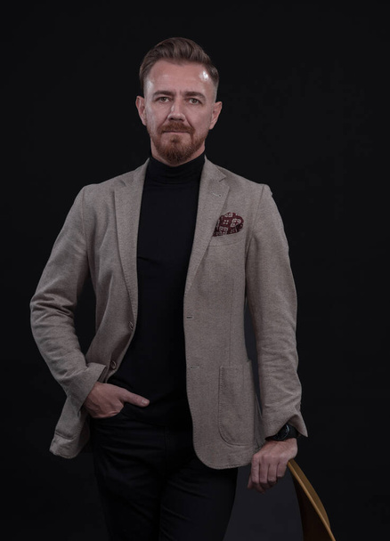 Portrait of adult businessman wearing trendy suit and sitting in modern studio on stylish chair against the black background. Horizontal mockup. High quality photo - Valokuva, kuva