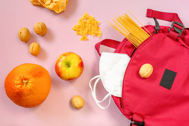 Красный рюкзак с различными макаронами, фруктами и орехами, защитная маска на розовом фоне. Концепция доставки еды, покупки, туристическая еда. Вид сверху - Фото, изображение