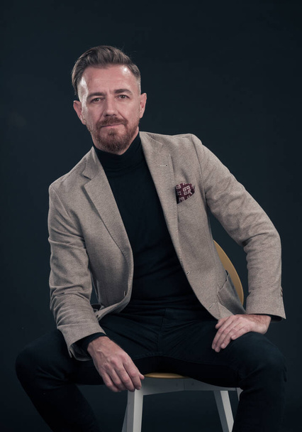 Portrait of adult businessman wearing trendy suit and sitting in modern studio on stylish chair against the black background. Horizontal mockup. High quality photo - Фото, зображення