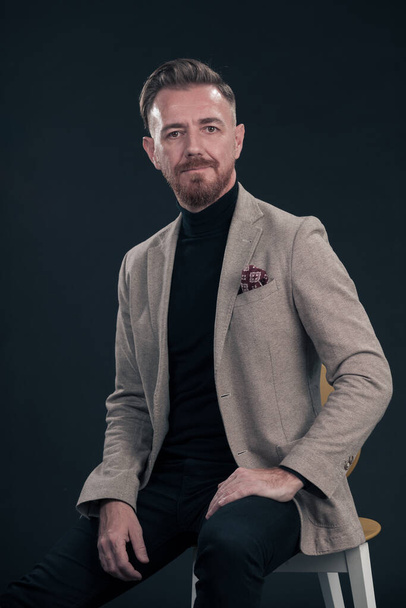 Portrait of adult businessman wearing trendy suit and sitting in modern studio on stylish chair against the black background. Horizontal mockup. High quality photo - Foto, immagini