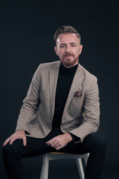 Portrait of adult businessman wearing trendy suit and sitting in modern studio on stylish chair against the black background. Horizontal mockup. High quality photo - Foto, Imagem