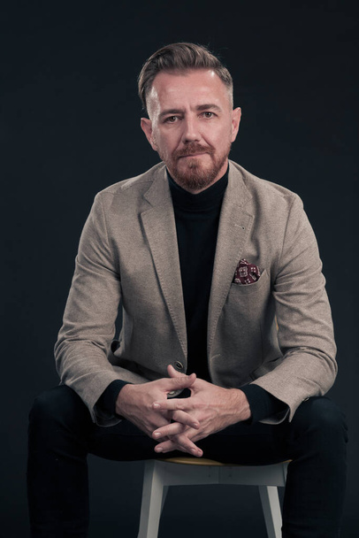 Portrait of adult businessman wearing trendy suit and sitting in modern studio on stylish chair against the black background. Horizontal mockup. High quality photo - Foto, Imagen