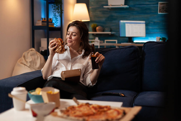 Smiling woman sitting on couch looking at tasty takeaway burger holding beer bottle in front of table witht pizza - Foto, Imagen