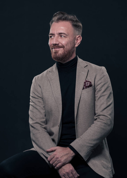 Portrait of adult businessman wearing trendy suit and sitting in modern studio on stylish chair against the black background. Horizontal mockup. High quality photo - Zdjęcie, obraz