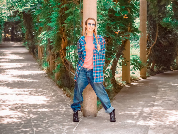 Possu nainen ruudullinen pitkä paita ylisuuret farkut ja karkea saappaat seisoo divarication puistossa nojaten pilariin - Valokuva, kuva