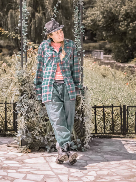 A girl in a hat and a careless oversized outfit is leaning on a green stand in a summer park - Foto, afbeelding