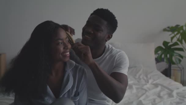 Szerelem és törődés. Boldog fiatal afro-amerikai férfi fésülködő haj barátnő, ül együtt a hálószobában, nyomkövető lövés - Felvétel, videó