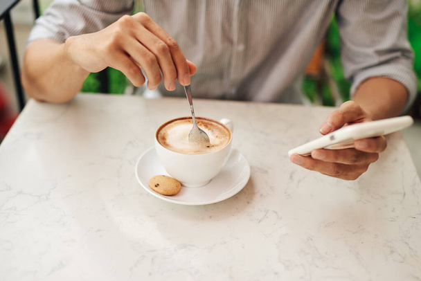 A man drinks cappuccino with a smartphone in his hand in a cozy restaurant - Photo, Image