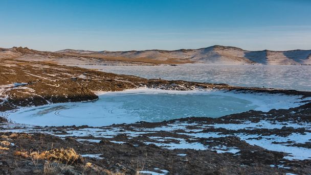 Pieni jäätynyt järvi sydämen muotoinen ja sen vieressä on suuri. Lunta ja kuivaa ruohoa maassa. Vuorijono sinistä taivasta vasten. Baikal. - Valokuva, kuva
