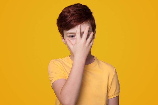 Muotokuva poika yllään rento t-paita seisoo yli eristetty keltainen tausta peittää osan kasvojen käsin. Eleitä. Lapsuus - Valokuva, kuva