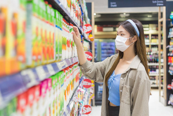 黄金のブロンドの長い髪を持つ若いアジアの女性は、彼女が新しい通常の概念でCOVID-19を保護するために医療面マスクを身に着けている間、週末にスーパーマーケットで商品を買い物に歩いています. - 写真・画像