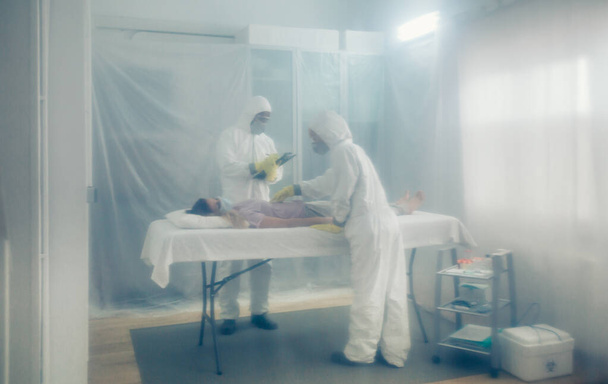 Bakteriyolojik koruma altındaki doktorlar sedyede yatan hastaya tıbbi muayene yapıyorlar. - Fotoğraf, Görsel
