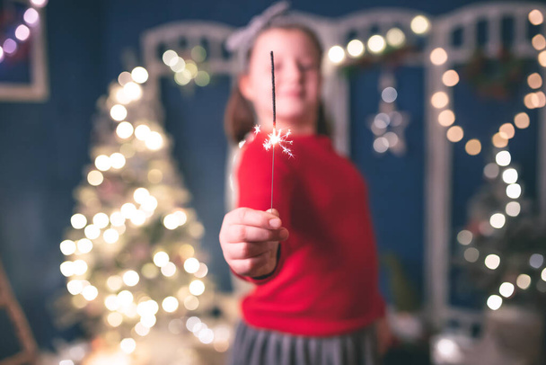 Young girl holding burning sparkler on festive holiday lights background. Christmas holidays, childhood and people concept - Foto, immagini