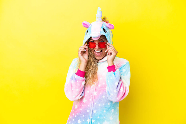 Girl with curly hair wearing a unicorn pajama isolated on yellow background with glasses and surprised - Foto, afbeelding