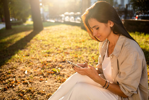 retrato de una hermosa mujer usando el teléfono en la hierba al aire libre con una vista de la ciudad al atardecer. - Foto, Imagen