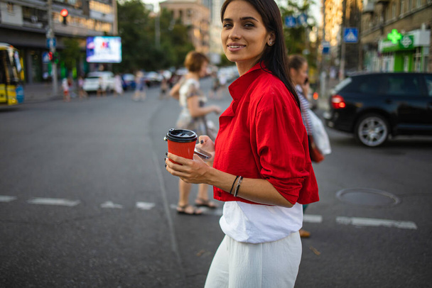Close-up vrouwelijke handen houden smartphone en koffie op de achtergrond van de straat crosswalk. Jonge zakenvrouw met behulp van mobiele apparaat staan op de weg stad - Foto, afbeelding