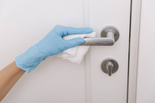 Cleaning black door handle with an antiseptic wet wipe in blue gloves. Woman hand using towel for cleaning. Sanitize surfaces prevention in hospital and public spaces against corona virus - Photo, image