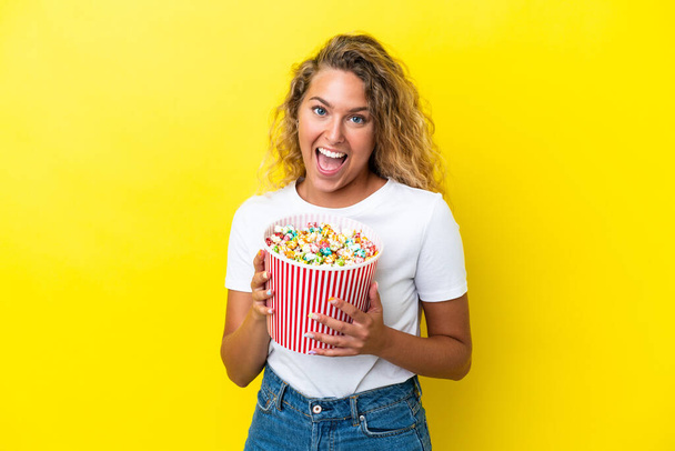 Girl with curly hair isolated on yellow background holding a big bucket of popcorns - Photo, Image