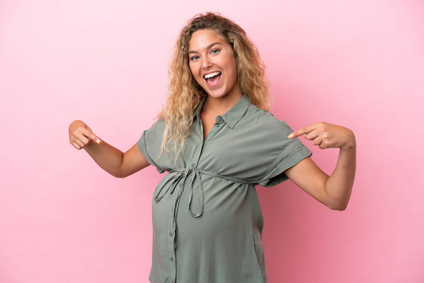 Girl with curly hair isolated on pink background pregnant and pointing to the belly - Fotoğraf, Görsel