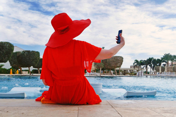 Vacances d'été femme en chapeau rouge et cape pour la plage avec téléphone mobile près de la piscine et palmiers amd, femme blogueuse, en plein air - Photo, image