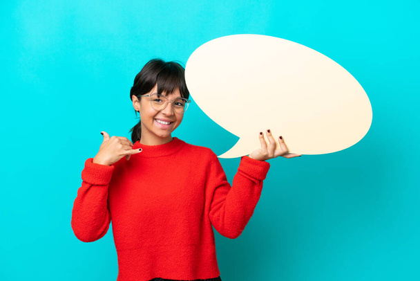 Young woman isolated on blue background holding an empty speech bubble and doing phone gesture - Photo, Image