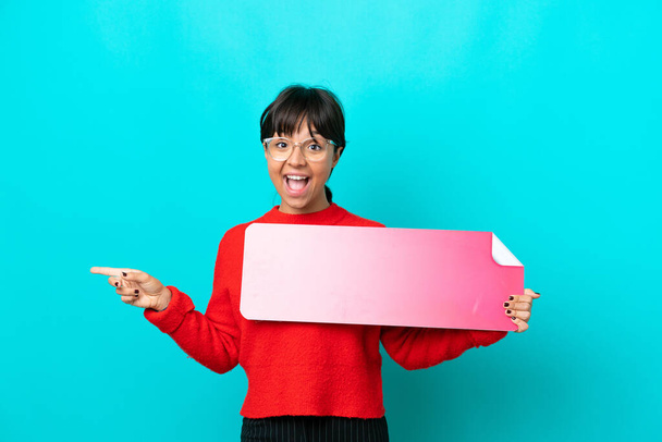 Young woman isolated on blue background holding an empty placard and pointing side - Фото, изображение