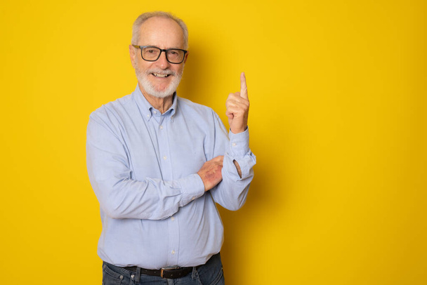Старший сіро-волохатий чоловік в елегантній сорочці стоїть над ізольованим жовтим тлом, показуючи і вказуючи пальцем номер один, посміхаючись впевнено і щасливо
. - Фото, зображення