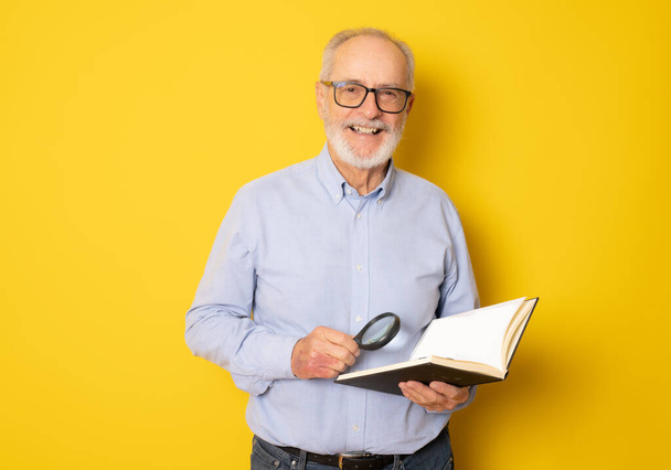 Χαμογελώντας ηλικιωμένος διαβάζοντας ένα βιβλίο απομονωμένο σε κίτρινο φόντο. - Φωτογραφία, εικόνα