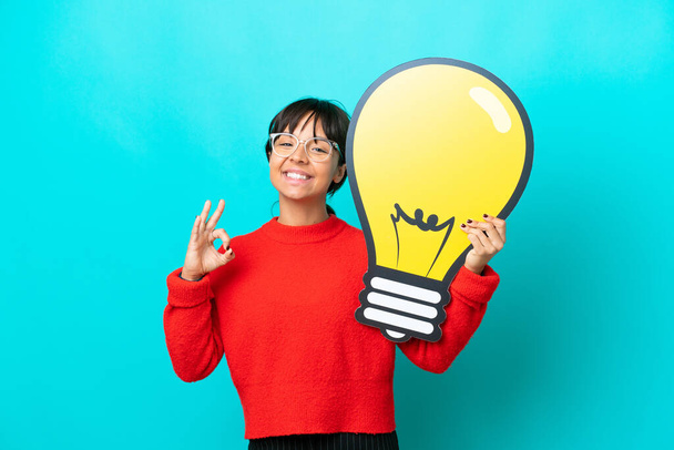 Young woman isolated on blue background holding a bulb icon and doing OK sign - Photo, Image