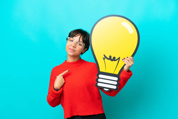 Young woman isolated on blue background holding a bulb icon and pointing it - Photo, Image
