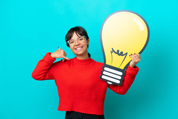 Young woman isolated on blue background holding a bulb icon and doing phone gesture - Photo, Image