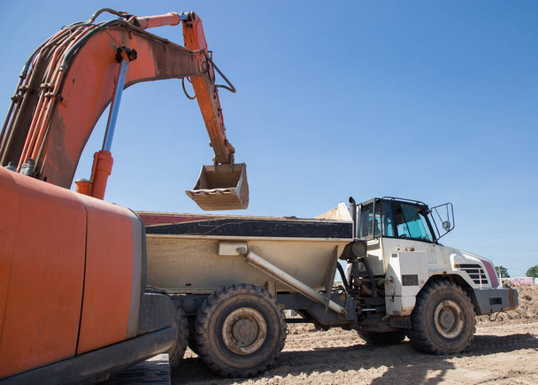 excavadora de orugas anaranjadas y un volquete de construcción articulado durante la excavación en un pozo de arena en un día soleado caliente. carga y transporte del suelo - Foto, Imagen