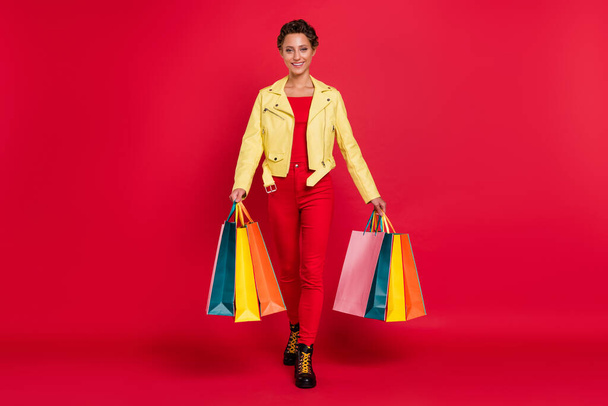 Full body photo of charming pretty happy woman walk hold hands many shopper bags smile isolated on red color background - 写真・画像