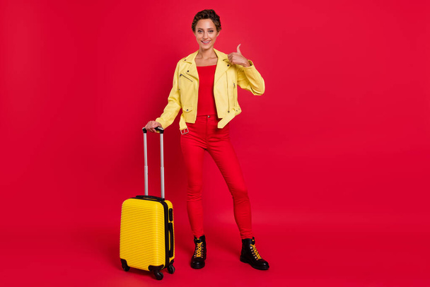 Fotografie lesklé sladké mladé ženy oblečené kožené bundy drží zavazadla ukazující palec nahoru s úsměvem izolované červené barvy pozadí - Fotografie, Obrázek