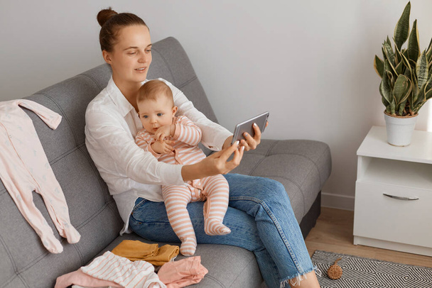 Caucasian female blogger with dark hair and bun hairstyle wearing casual clothing sitting on cough with toddler baby and shooting video for her moms vlog or having livestream. - Zdjęcie, obraz