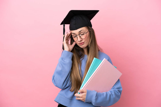 fiatal diák nő visel diplomás kalap elszigetelt rózsaszín háttér fejfájás - Fotó, kép