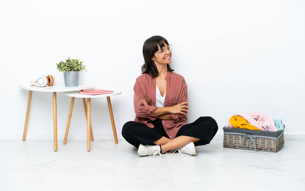 Young mixed race woman folding clothes sitting on the floor isolated on white background with arms crossed and looking forward - Фото, изображение