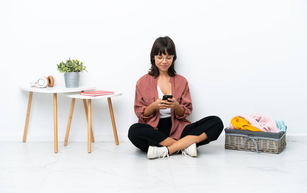 Junge Frau mit gemischter Rasse faltet Klamotten auf dem Boden sitzend isoliert auf weißem Hintergrund und sendet eine Nachricht mit dem Handy - Foto, Bild