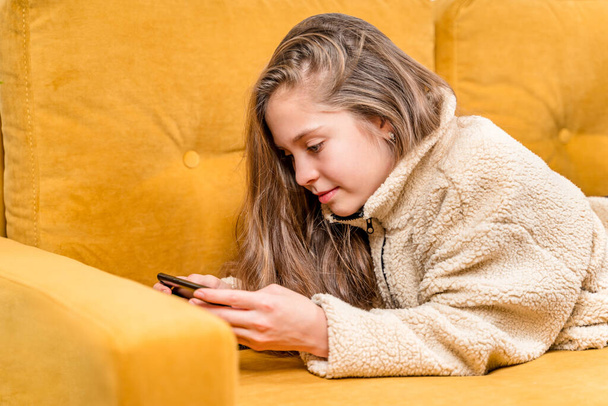 Kaunis pieni tyttö teini-iässä makaa keltaisella sohvalla ja leikkii matkapuhelimella tai kommunikoi ystävien kanssa, valikoiva keskittyminen. - Valokuva, kuva