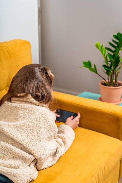 Une belle petite fille dans son adolescence se trouve sur un canapé jaune et joue avec un téléphone portable ou communique avec des amis, mise au point sélective - Photo, image
