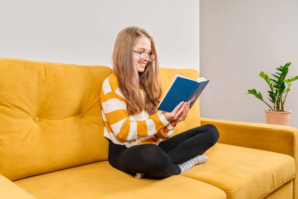Een klein tienermeisje met een bril zit thuis op een gele bank en leest een boek met een slimme grappige uitdrukking op haar gezicht. Leerconcept - Foto, afbeelding