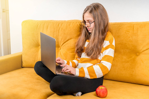 Sonriente adolescente europea está utilizando una computadora portátil para videollamadas a amigos y padres sentados en un sofá amarillo. Educación a distancia en línea. - Foto, imagen